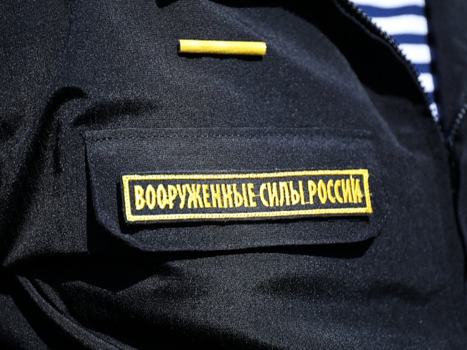На изображении чёрная ткань с жёлтой нашивкой, на которой написано "Вооружённые Силы России". Над нашивкой жёлтая пуговица..