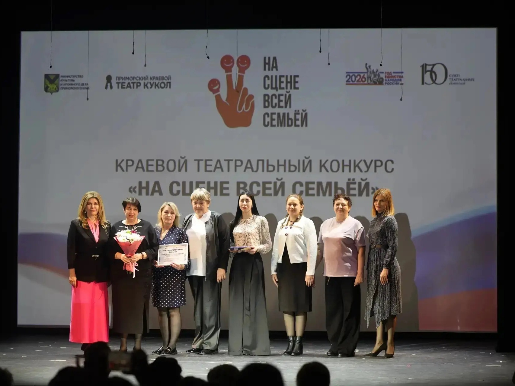 На сцене семь женщин, стоящих в ряд. На заднем плане экран с надписью «На сцене всей семьёй». Две женщины держат цветы и грамоты..