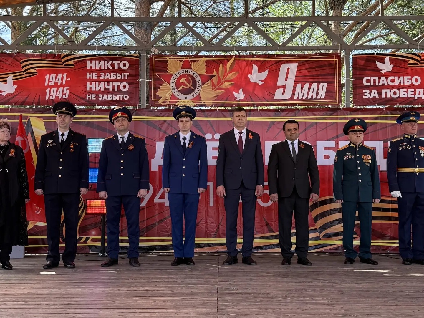 На изображении семь человек в форме стоят на сцене. На фоне красные баннеры с надписями о Великой Отечественной войне и 9 мая..