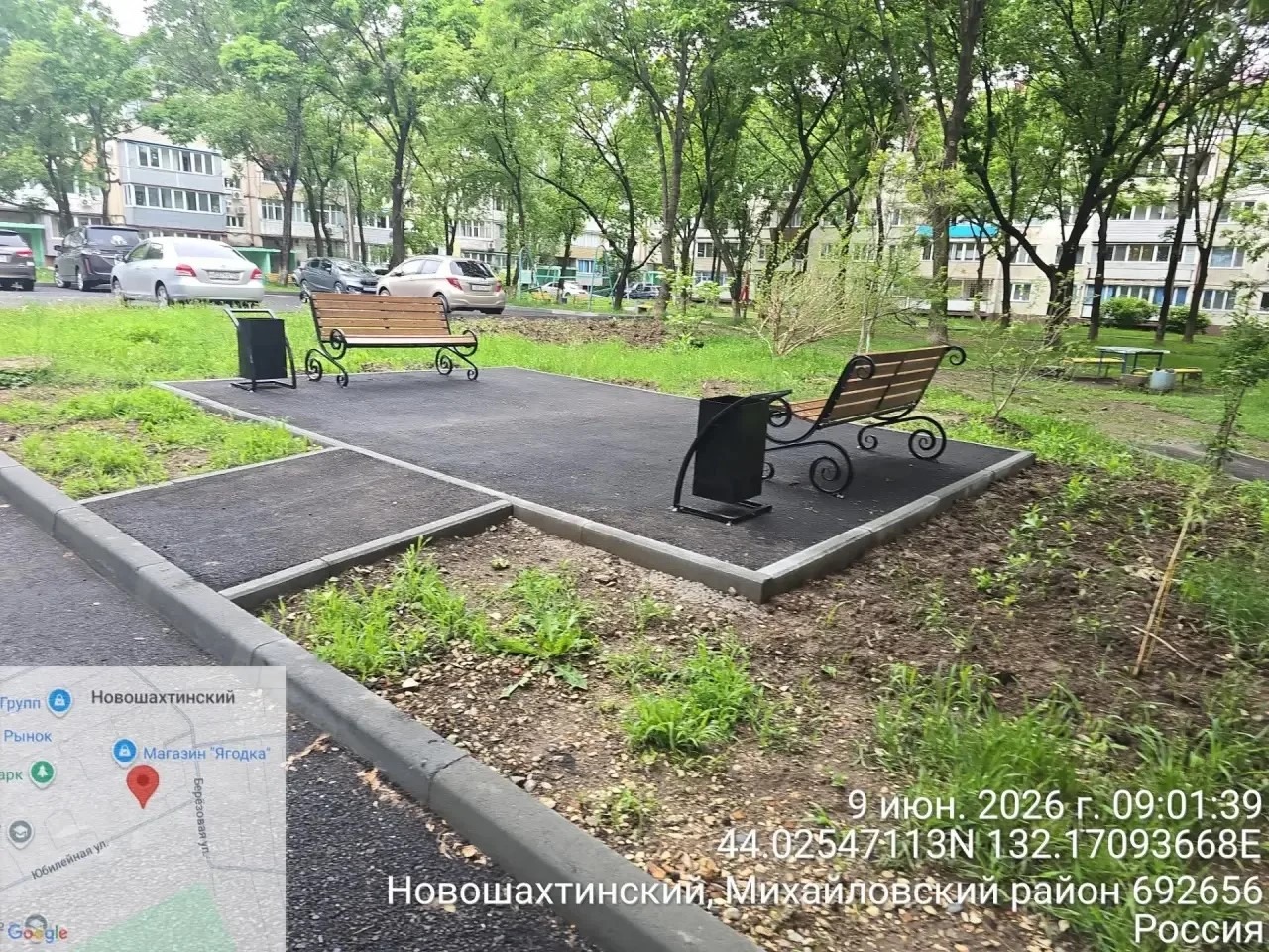 На изображении показана парковая зона с двумя скамейками, урнами и дорожками из асфальта. Вокруг растут деревья и зелёная трава.